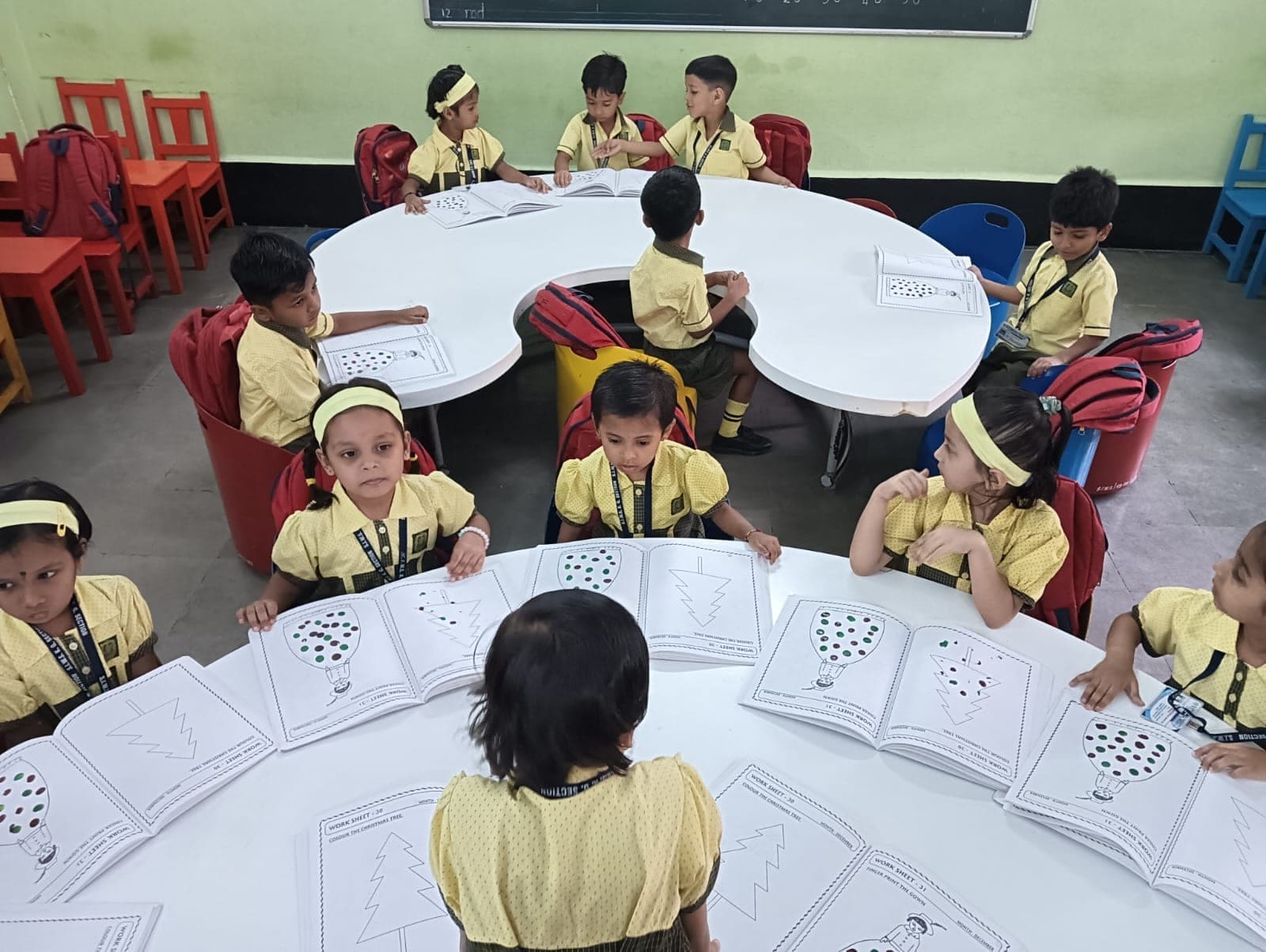 Kindergarten children seated at curved tables working through printed worksheets, with a number line on the blackboard behind them.