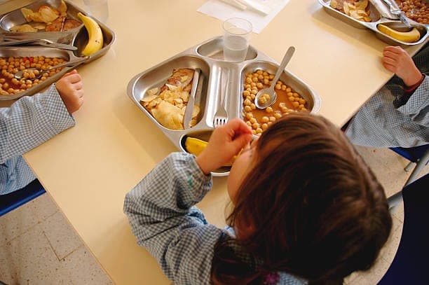 A young child at a dining table eating a school meal from a sectioned metal tray.