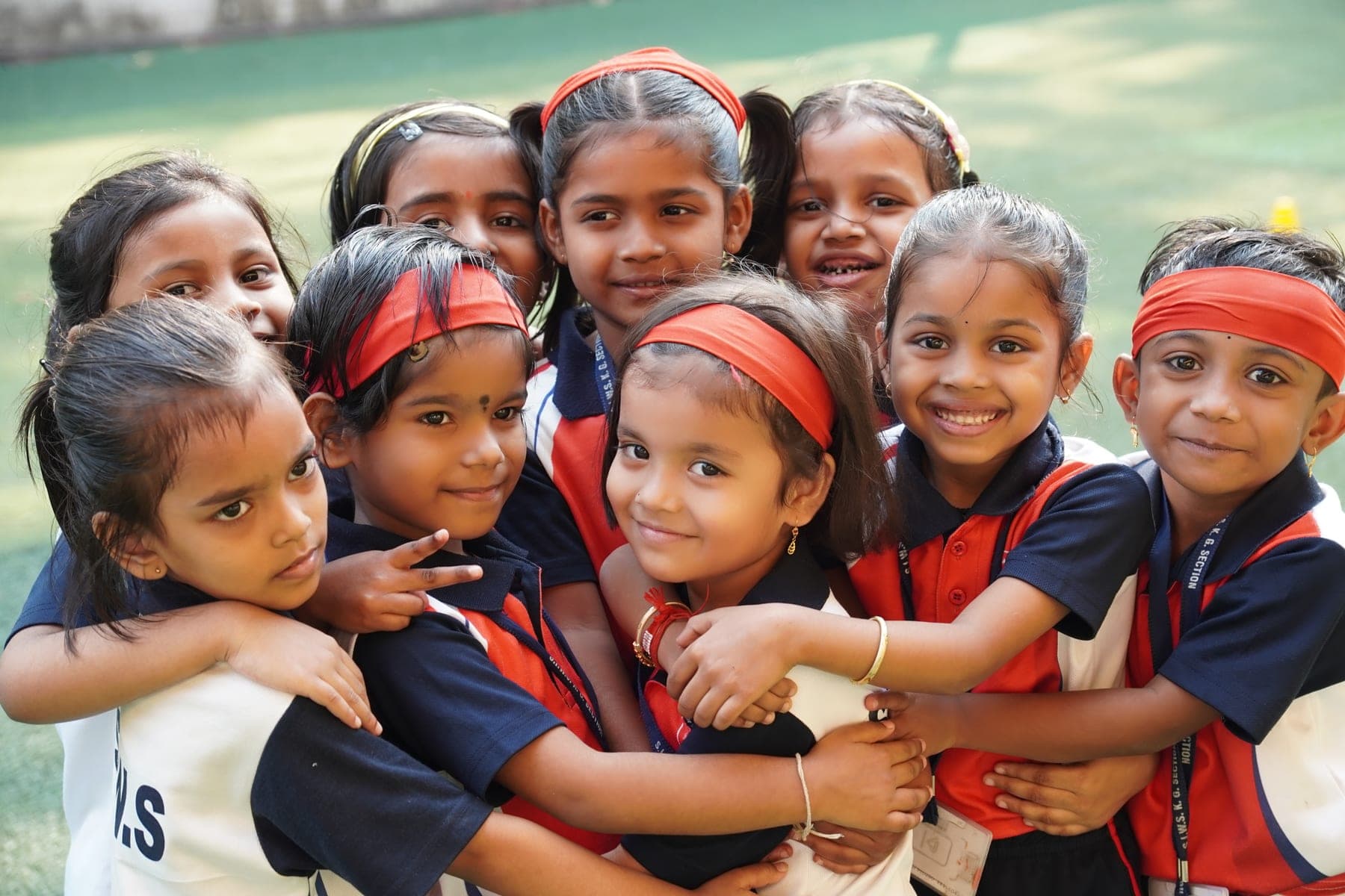 A close group of kindergarten girls in SIWS sports uniform with red headbands, arms around each other, smiling.