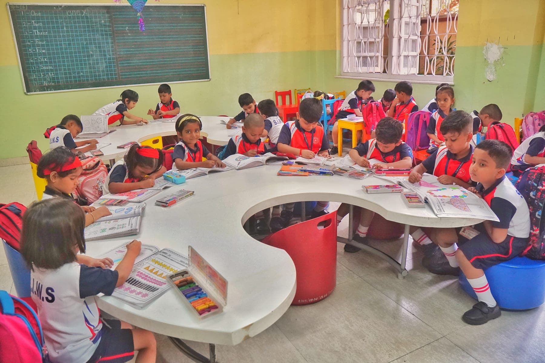 Kindergarten children in SIWS uniform sitting at curved group tables, colouring in activity books with crayons.