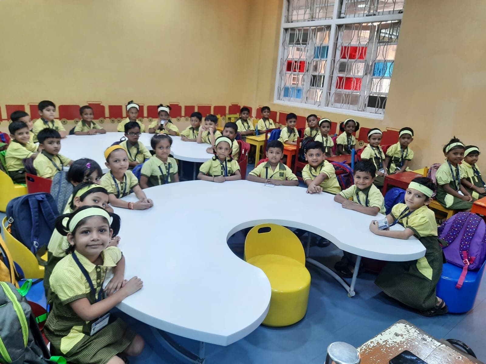 A full kindergarten class in yellow and green SIWS uniform, seated around two large curved white tables and turning to face the camera.