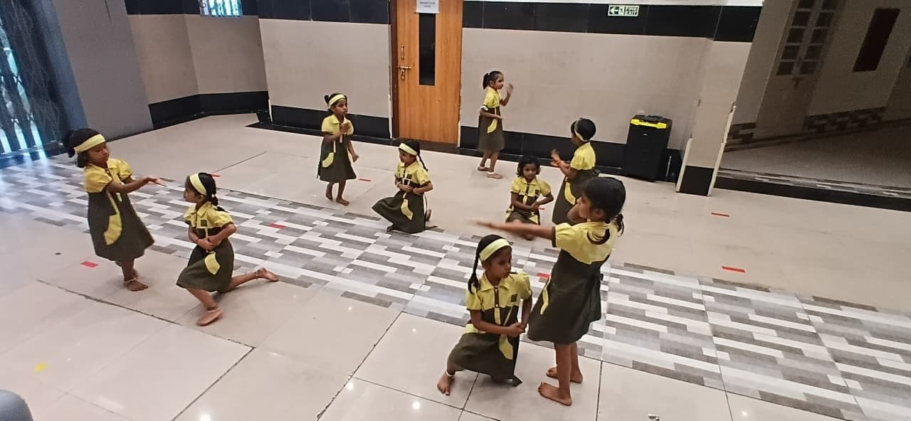 Kindergarten children rehearsing a dance in the school hall, barefoot and in school uniform with yellow headbands, spread across floor markings mid-step.