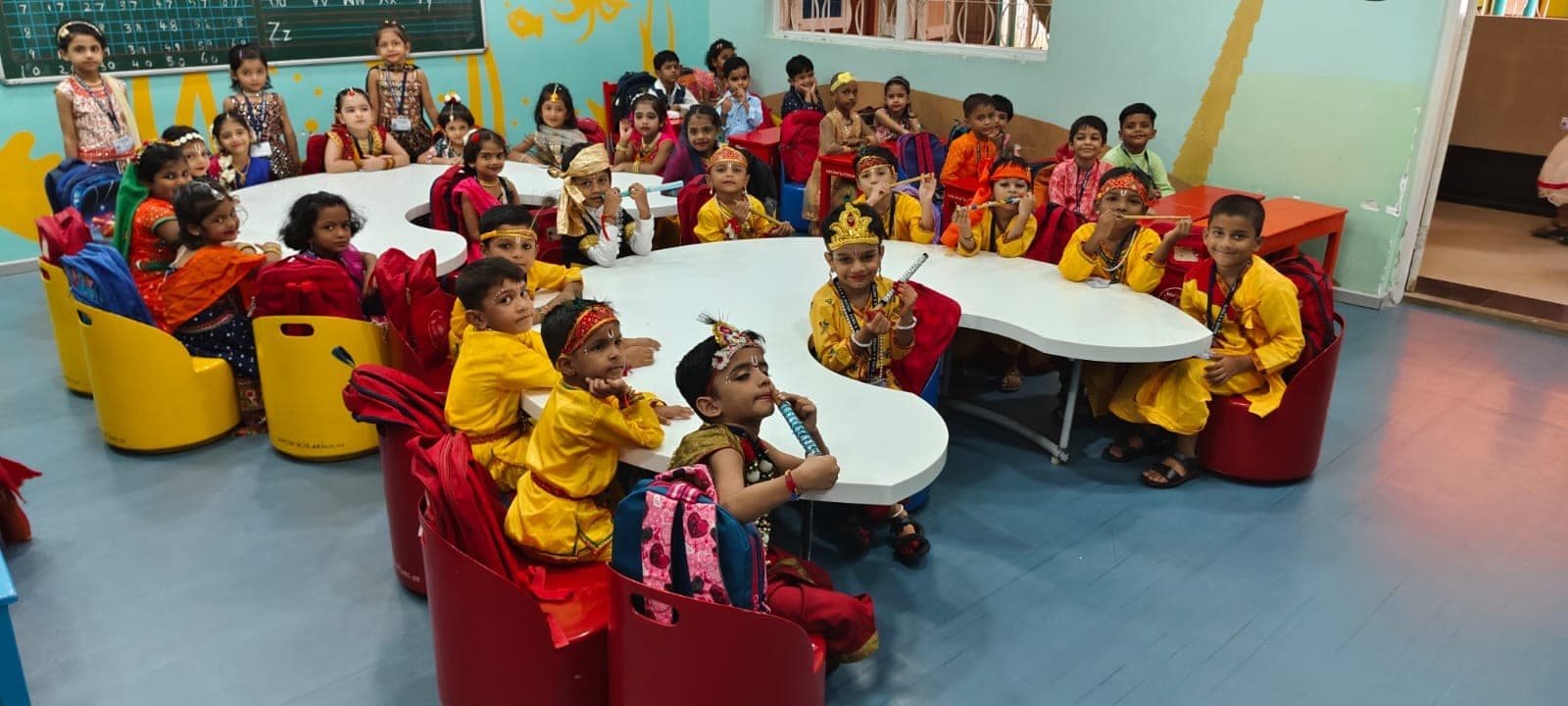 Kindergarten children dressed as Krishna and Radha at their classroom tables for Janmashtami, several holding flutes.