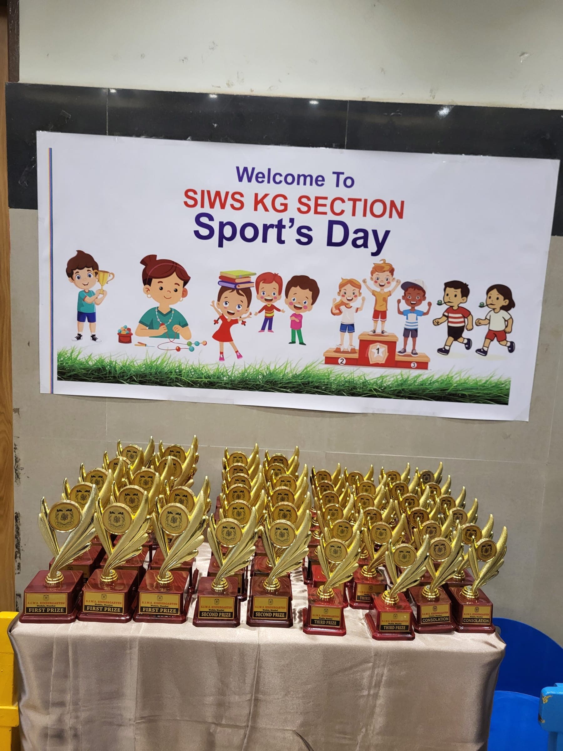 A table covered with rows of gold trophies labelled First Prize, Second Prize and Consolation, beneath a printed banner reading “Welcome To SIWS KG SECTION Sport’s Day”.