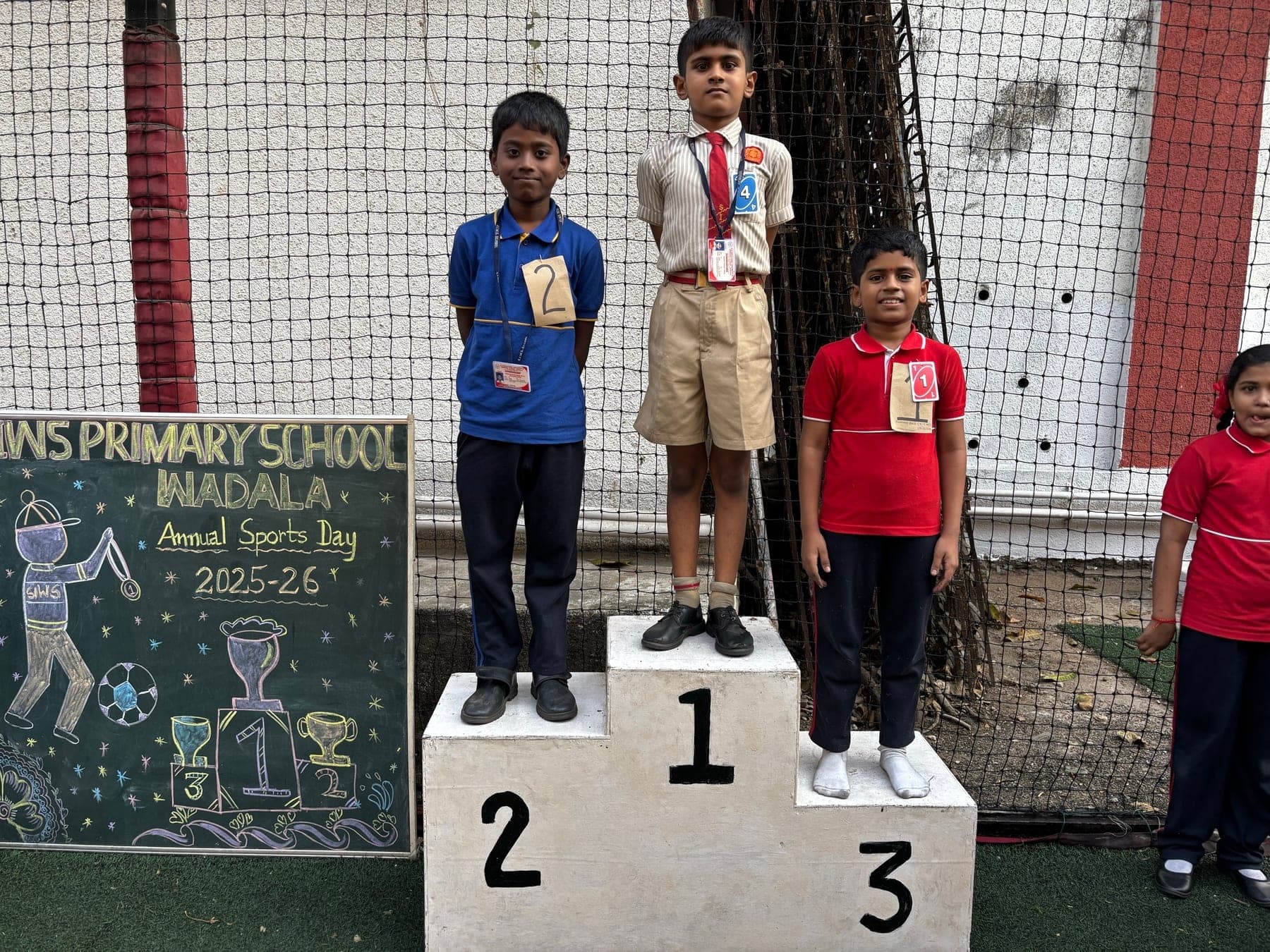 Three Primary boys on the winners’ podium — second in blue, first in a cream shirt, third in red — beside the Annual Sports Day 2025-26 chalkboard.