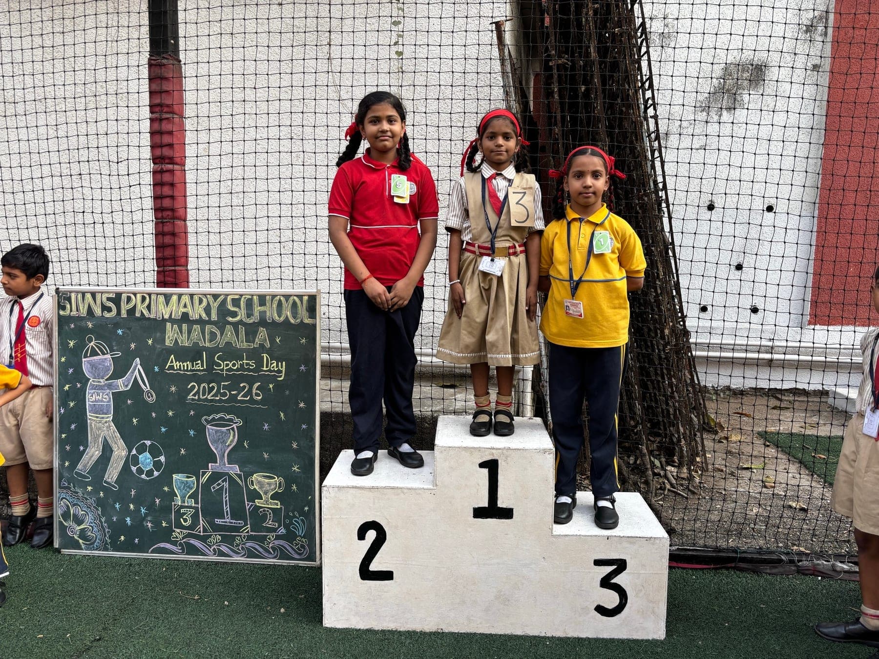 Three Primary girls on the winners’ podium — second in red, first in a cream pinafore with a red headband, third in yellow — beside the Annual Sports Day 2025-26 chalkboard.
