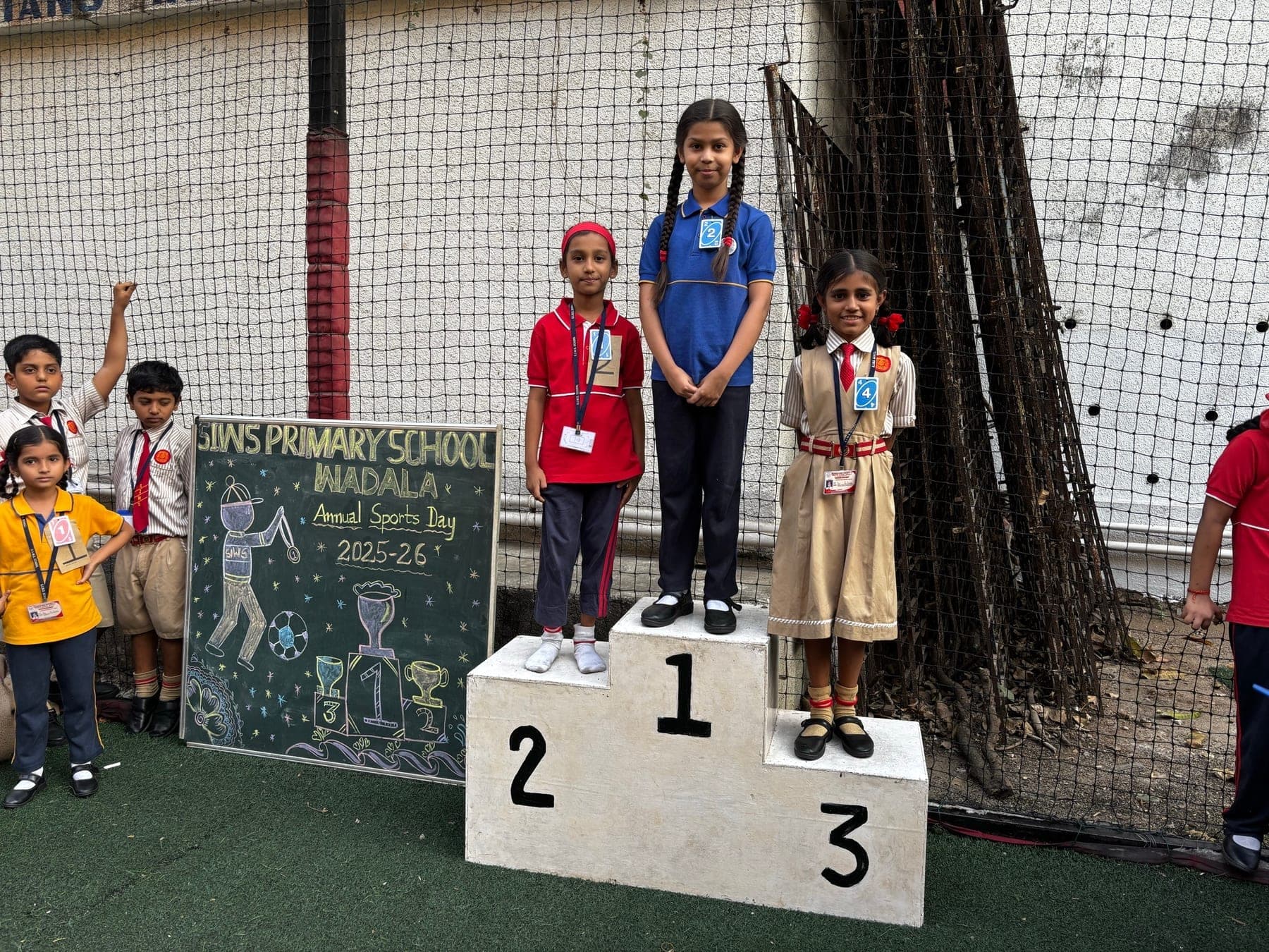 Three Primary girls on the winners’ podium — second in red with a red headband, first in blue, third in a cream pinafore — with classmates waiting at the edge of the turf pitch.