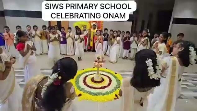 Children in cream Kerala dress standing with hands folded around a circular flower pookalam and a lit lamp, under a caption reading “SIWS Primary School celebrating Onam”.