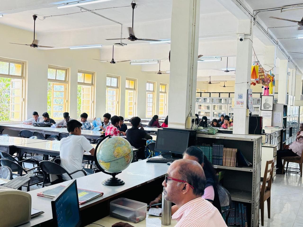 The college library on an ordinary afternoon — students reading and working at long tables by a row of windows, with book stacks, computers and a globe on the counter.