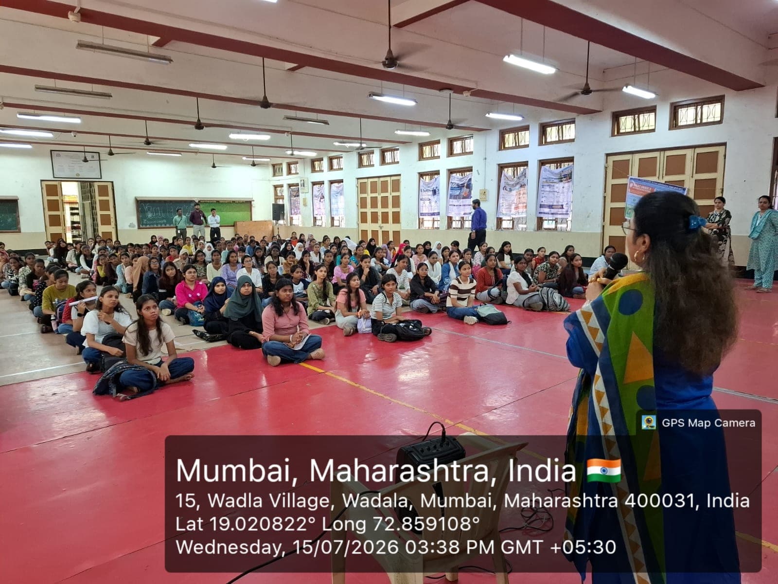 The college hall filled with seated students listening to a speaker addressing them with a microphone, with staff standing along the sides.