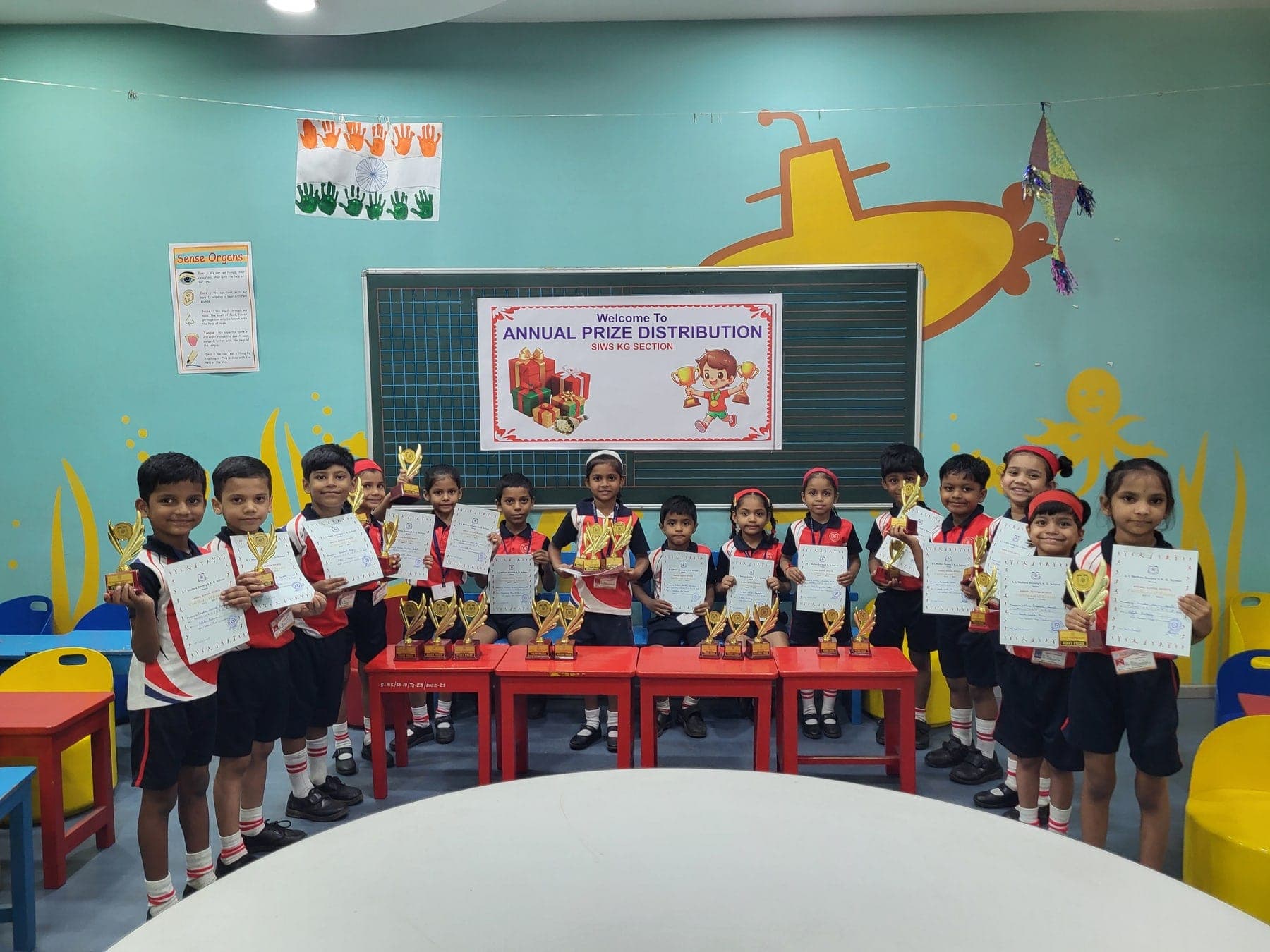 Fifteen Kindergarten children in red and black sports kit holding trophies and Annual School Sports certificates, in a classroom painted with a yellow submarine, beneath a banner reading “Annual Prize Distribution — SIWS KG Section”.