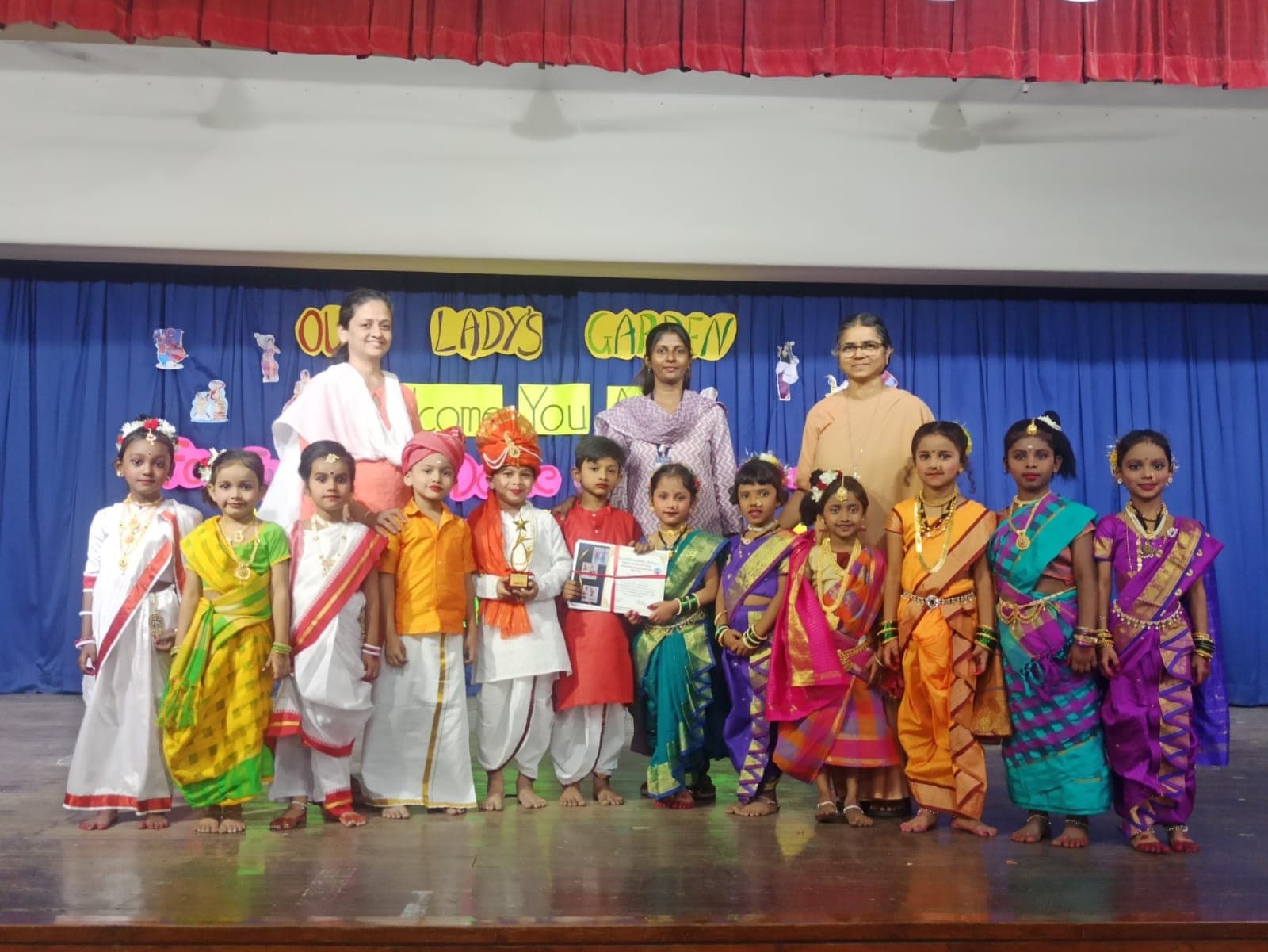 Thirteen Kindergarten children in regional Indian costume on stage with three teachers, one child holding a trophy and another a certificate, beneath an Our Lady’s Garden banner.