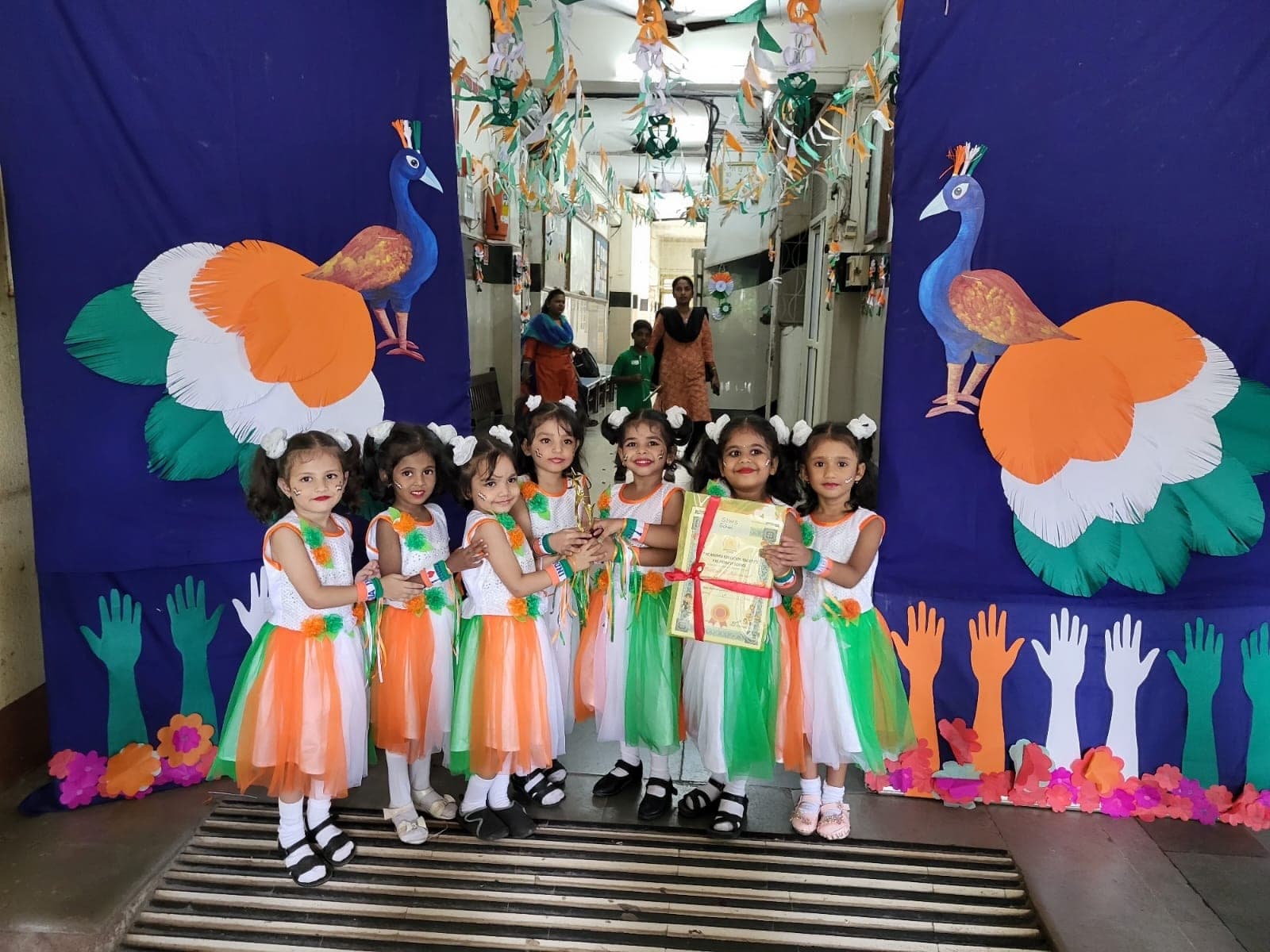 The same group of Kindergarten dancers in tricolour costume holding a trophy and a certificate from The Andhra Education Society’s Pre-Primary School, in a corridor decorated with paper peacocks.