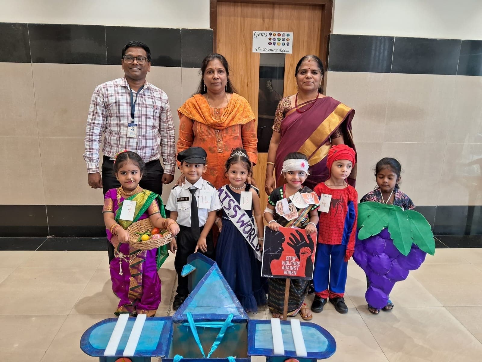 Six Kindergarten children in fancy dress with numbered cards pinned to them — a fruit seller, a pilot, a beauty queen, a campaigner, Spider-Man and a bunch of grapes — with three teachers behind and a cardboard aeroplane at their feet.