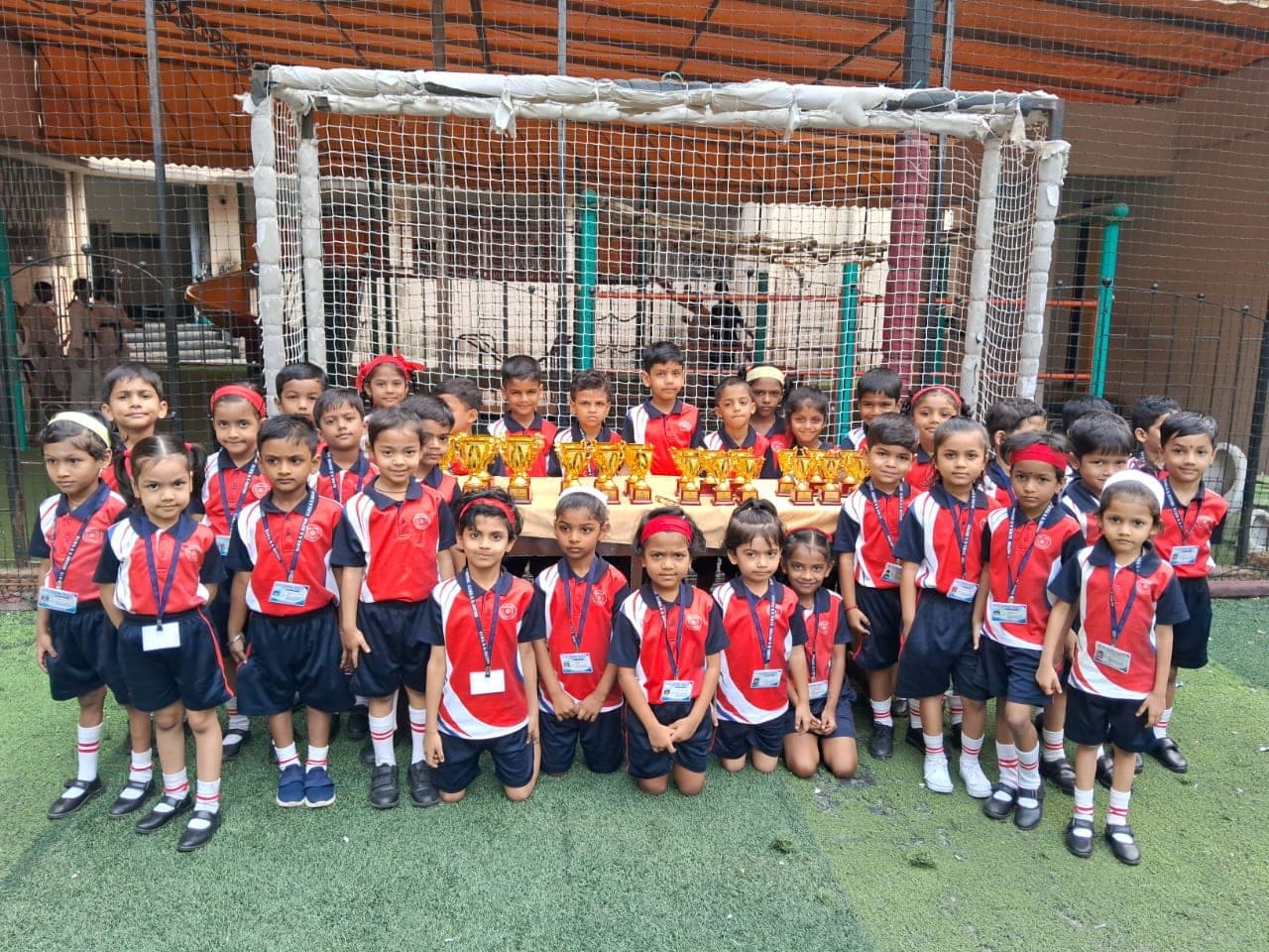 Around thirty Kindergarten children in red and navy sports kit standing in rows on the turf pitch in front of the goal, behind a table holding six gold trophies.