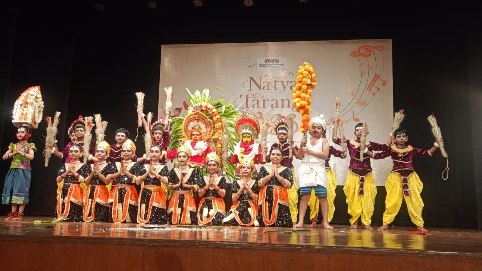 The full company of child dancers in costume and face paint on stage, some kneeling with palms together, others standing with decorated staffs.
