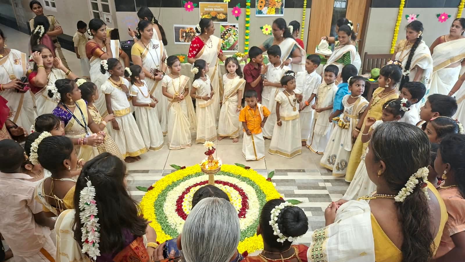 Children in cream-and-gold Kerala dress crowded around a large circular flower pookalam of yellow, white, green and red petals, beside a table of Kerala snacks.