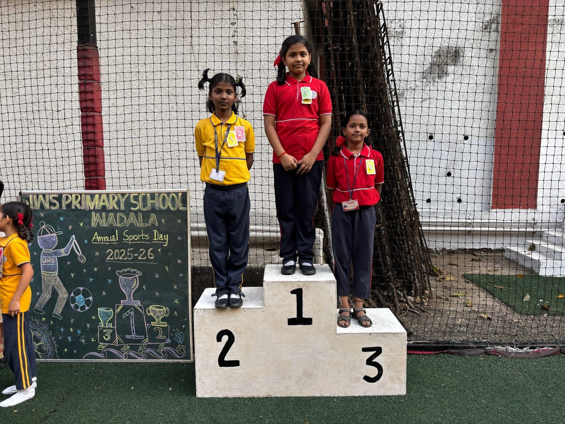 Three Primary girls on the winners’ podium — second in a yellow house shirt, first in red, third in red — beside a chalkboard reading “SIWS Primary School Wadala, Annual Sports Day 2025-26”.