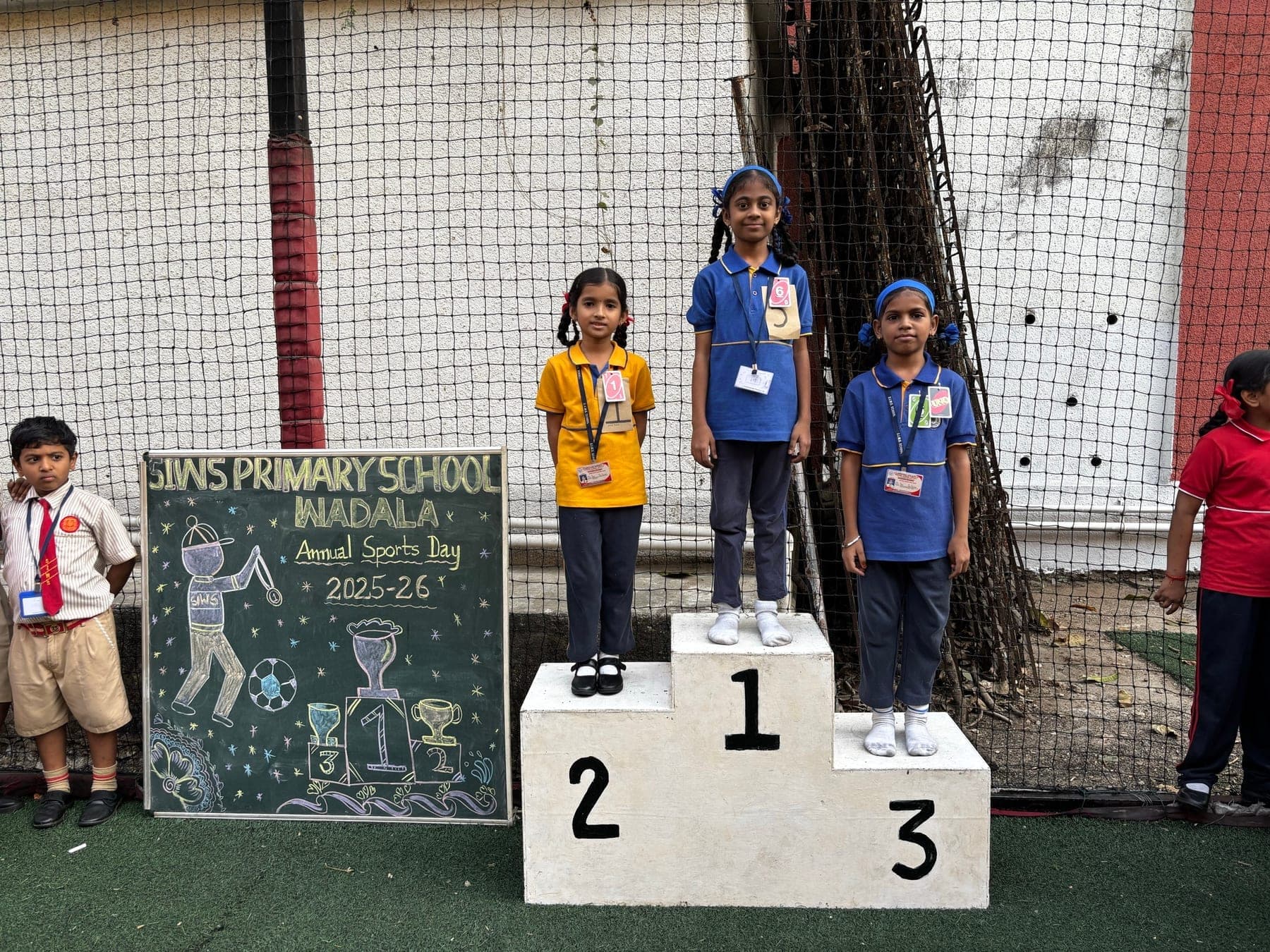 Three Primary girls on the winners’ podium — second in yellow, first in blue, third in blue — beside the Annual Sports Day 2025-26 chalkboard.