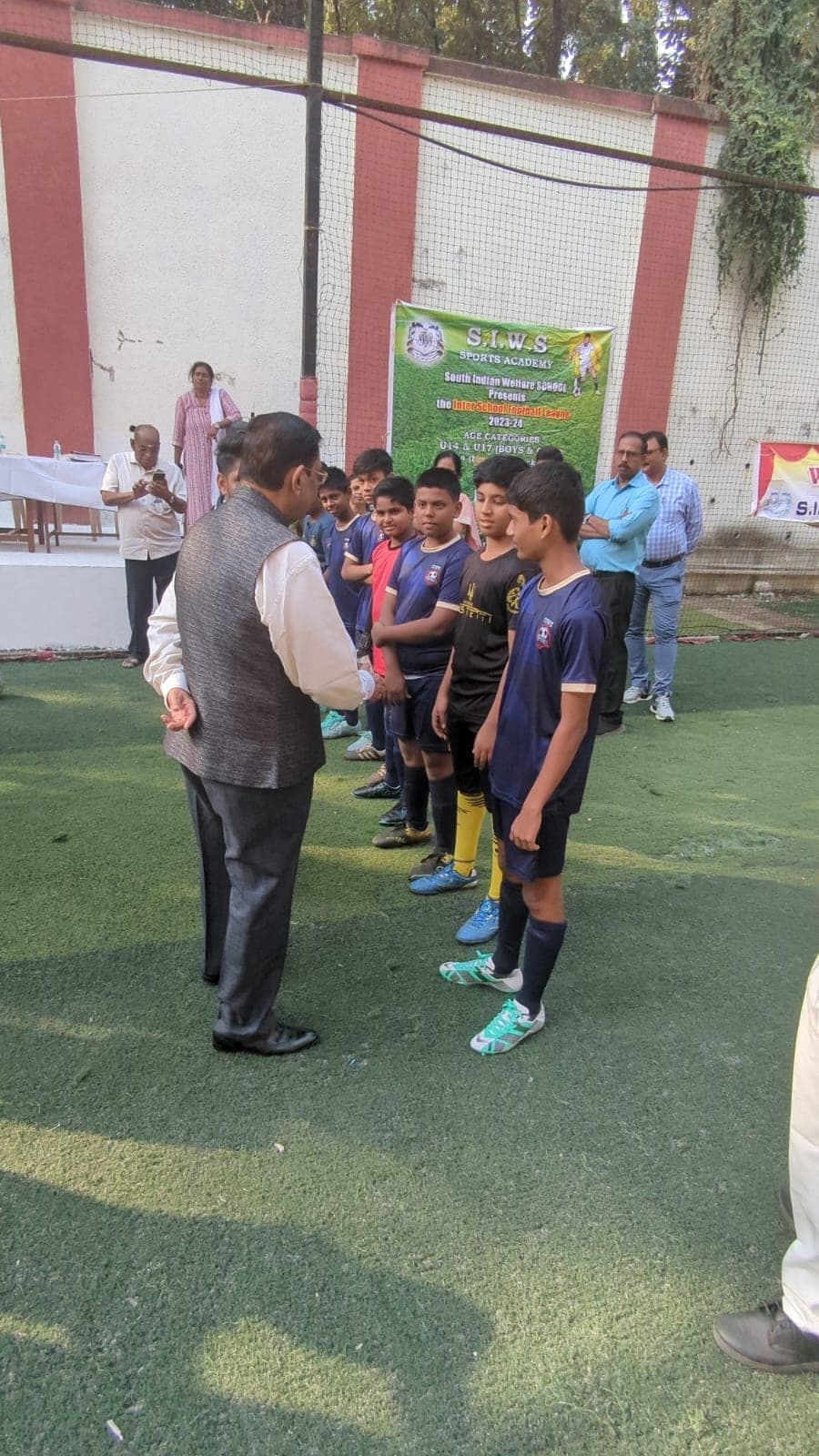 A line of boys in navy and yellow football kit standing on the turf pitch as a guest in a waistcoat greets them, in front of a banner for the S.I.W.S. Sports Academy Inter School Football League 2023-24, under-14 and under-17.