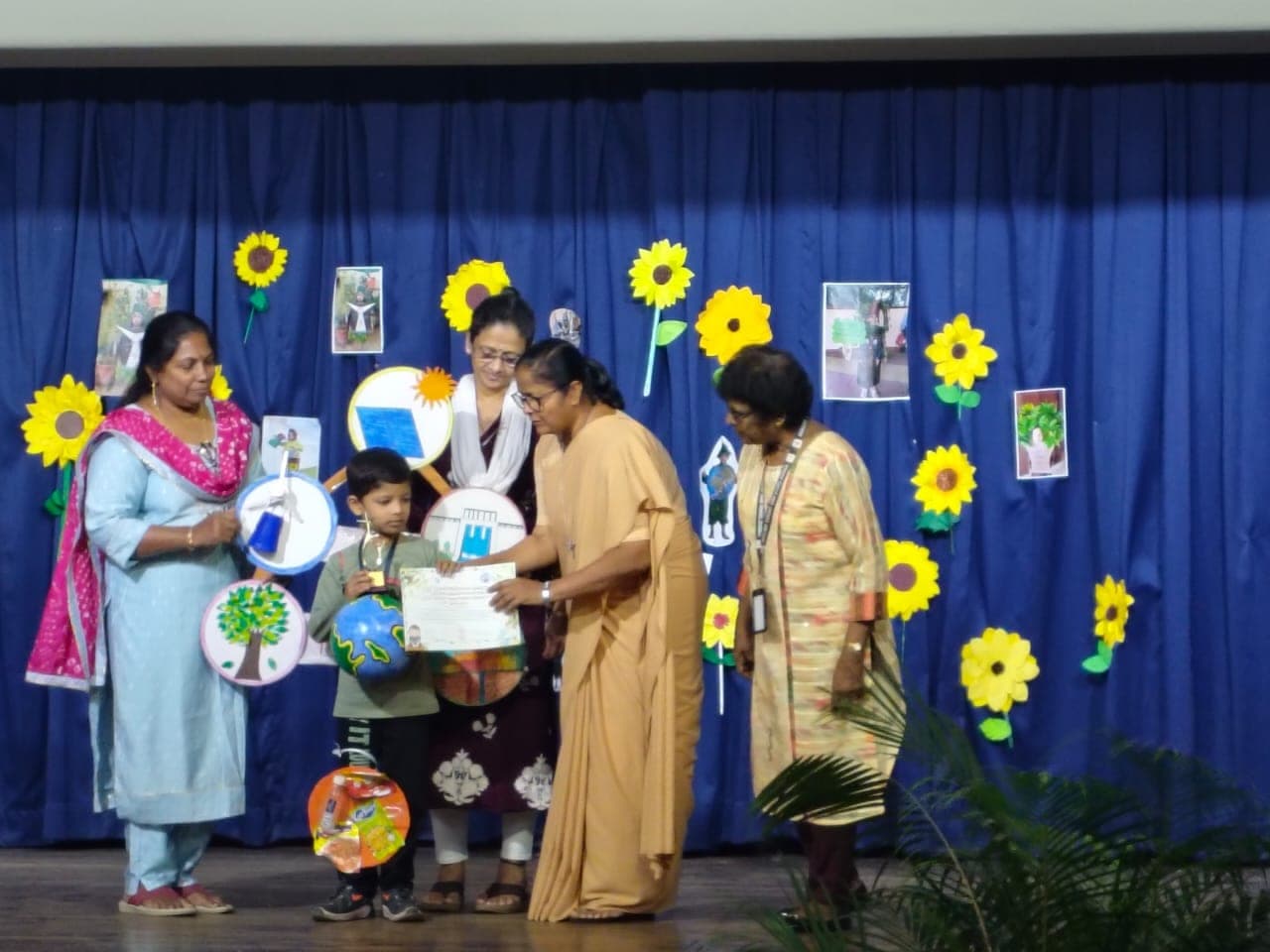 A kindergarten student in costume being handed a certificate on stage by a teacher, with three staff members alongside and a sunflower backdrop behind.