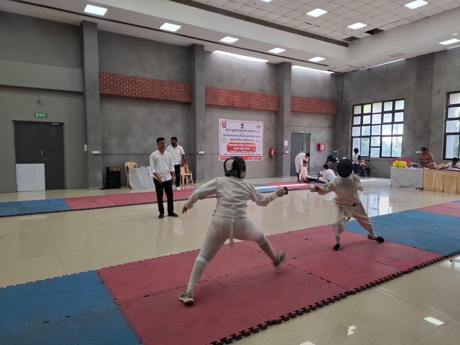 Two fencers in white kit and masks in the middle of a bout, one lunging, on a red and blue matted piste in a sports hall, with officials watching from the side.
