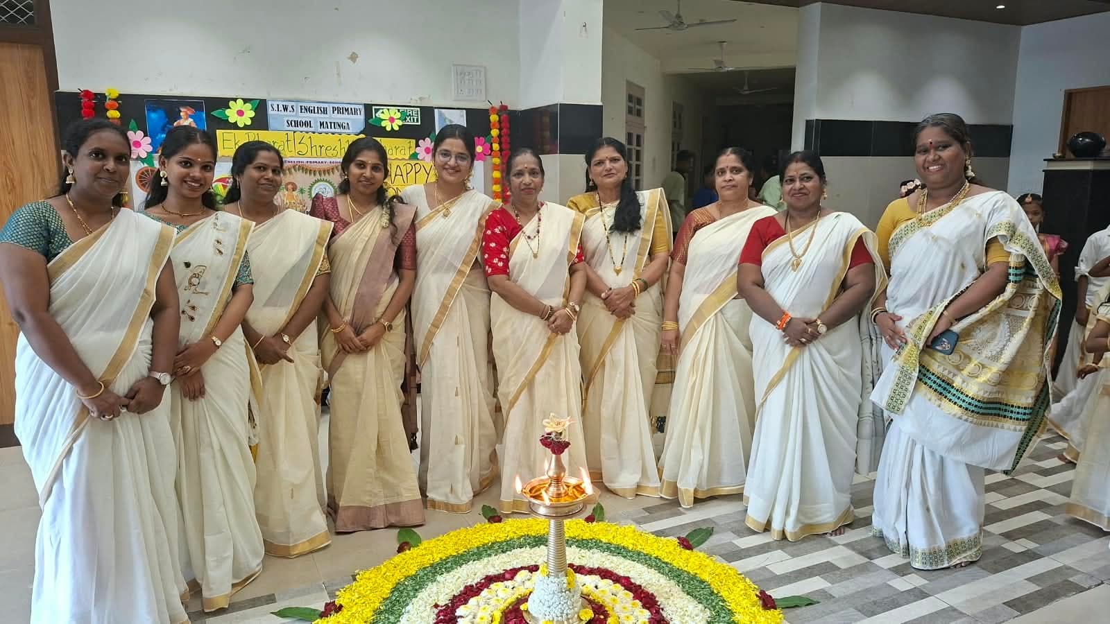 Ten members of staff in cream-and-gold Kerala sarees standing in a line behind the pookalam and lit lamp, in front of an Ek Bharat Shreshtha Bharat and Happy Onam display.