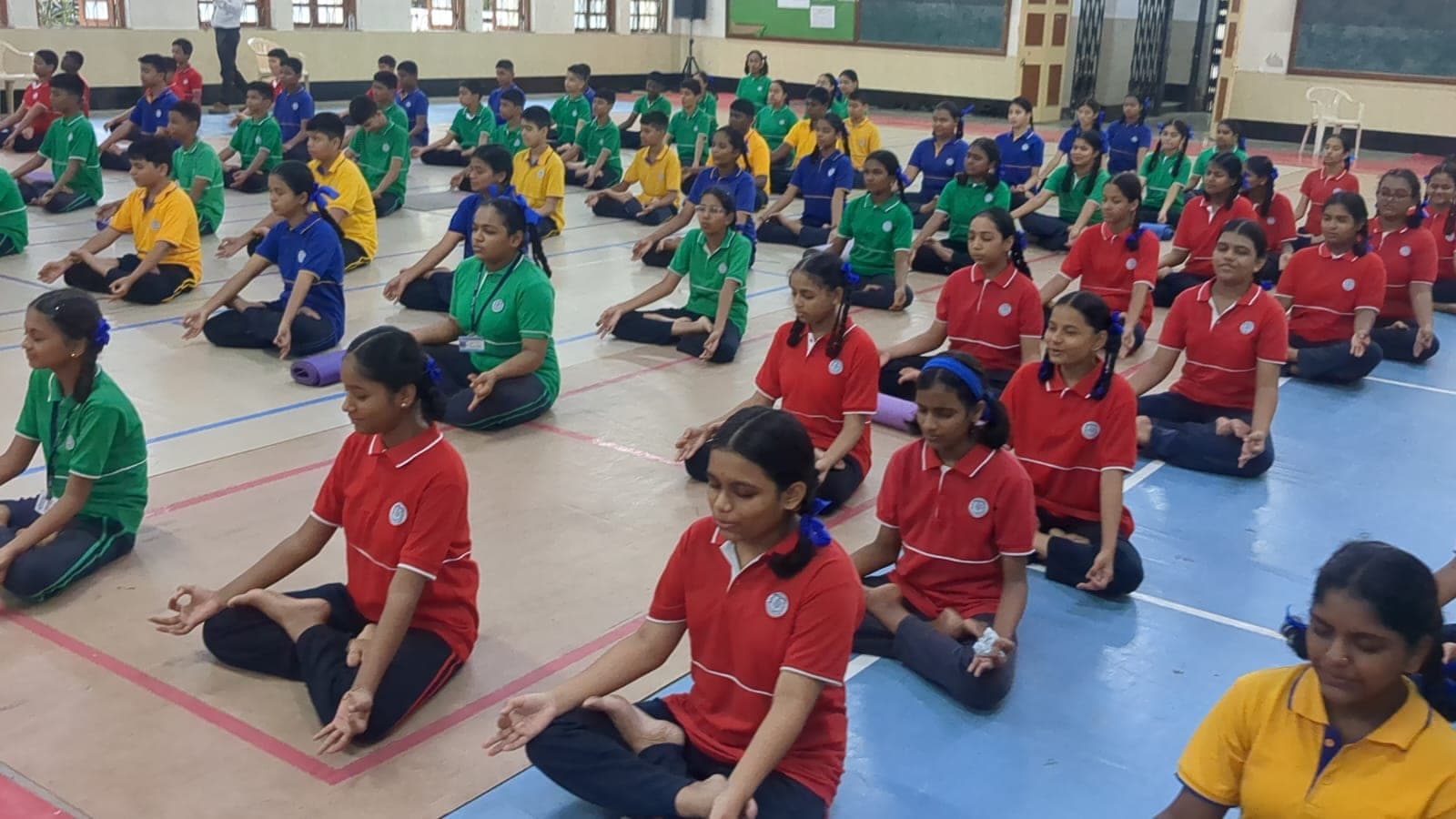 Rows of Secondary students in house-colour sports shirts seated cross-legged on mats in the school hall, eyes closed, during a guided meditation session.
